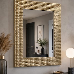 Pode incluir: Um espelho de parede retangular com uma moldura dourada larga e texturizada com um padrão de grade repetitivo. A superfície de vidro reflexiva mostra uma visão clara de um ambiente interno, incluindo uma porta escura, um quadro e um console.