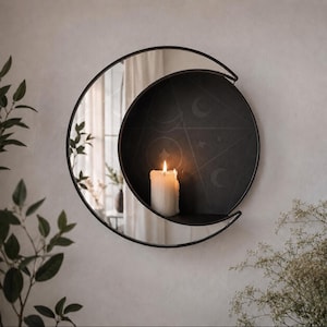 May include: A circular wall mirror with a black metal frame shaped like a crescent moon. The mirror features an integrated black shelf holding a white pillar candle. The shelf surface displays faint etched designs of stars and moon phases.