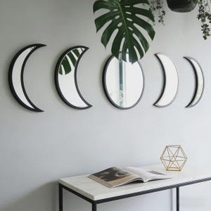 May include: A set of five wall-mounted mirrors featuring black frames. The arrangement includes two crescent moon shapes on the left, a central circular mirror, and two smaller crescent moon shapes on the right, reflecting a green plant leaf.