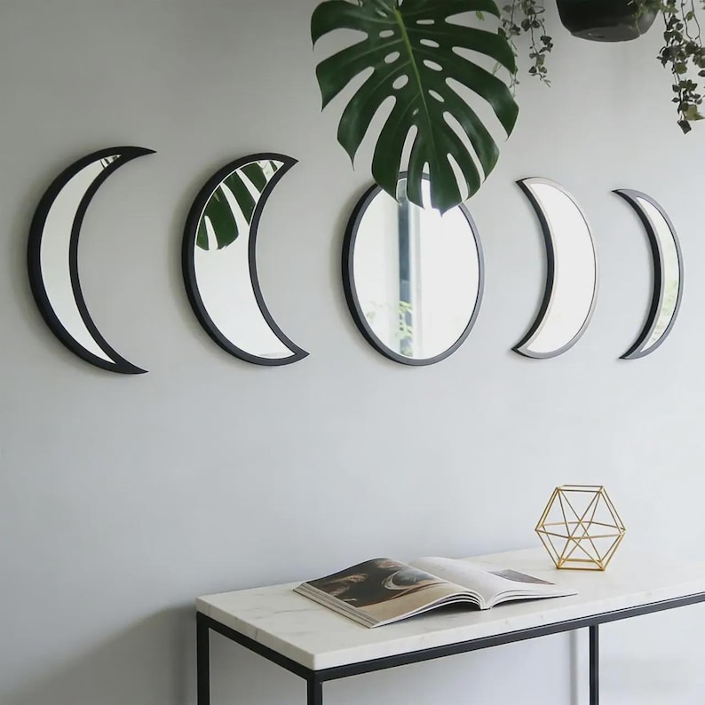 May include: A set of five wall-mounted mirrors featuring black frames. The arrangement includes two crescent moon shapes on the left, a central circular mirror, and two smaller crescent moon shapes on the right, reflecting a green plant leaf.