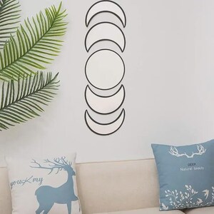 May include: A vertical wall-mounted mirror set featuring five distinct pieces arranged to represent the phases of the moon. The set includes one central circular mirror flanked by four crescent-shaped mirrors with thin black frames, creating a linear display.