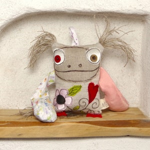 Peut inclure: Un jouet en peluche fait main avec un corps en tissu brun, des yeux en boutons rouges et des bras en tissu à motifs floraux et roses. Le jouet a une broderie en forme de cœur et de fleur sur le devant.