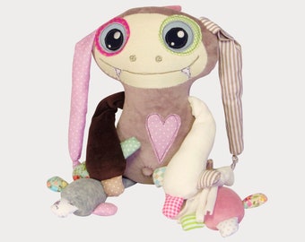 May include: A plush toy monster with brown fur, pink and white polka dot ears, and a pink heart on its chest. The monster has a toothy grin and large blue eyes with pink and green accents. The toy has a brown arm and a white arm with pink and green accents. The toy is sitting on a white background.