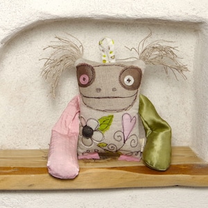 Peut inclure: Un jouet en peluche fait main et fantaisiste avec un corps en lin brun, des bras en tissu rose et vert et des détails brodés. Le jouet a de grands yeux bruns avec des boutons roses, un sourire cousu et un motif floral sur la poitrine.
