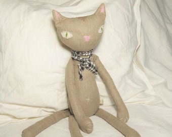 Puede incluir: Un juguete de peluche hecho a mano con forma de gato, con un cuerpo de lino gris, nariz rosa y ojos verdes. El gato lleva una bufanda a cuadros negros y blancos.