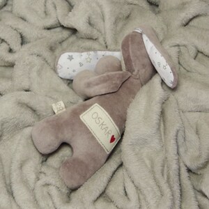 Puede incluir: Un conejito de peluche marr&oacute;n con una oreja blanca y un parche blanco en la espalda. El conejito tiene una etiqueta con el nombre "OSKAR" y un coraz&oacute;n rojo. El conejito est&aacute; acostado sobre una manta suave de color gris claro.