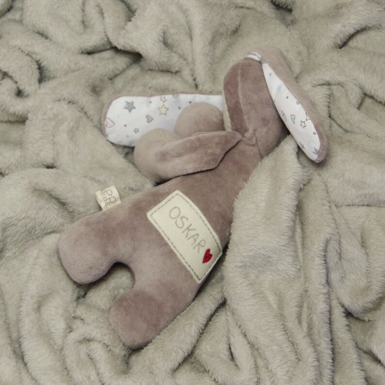 Puede incluir: Un conejito de peluche marr&oacute;n con una oreja blanca y un parche blanco en la espalda. El conejito tiene una etiqueta con el nombre "OSKAR" y un coraz&oacute;n rojo. El conejito est&aacute; acostado sobre una manta suave de color gris claro.