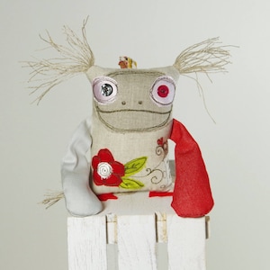 May include: A handmade plush toy with a gray linen body, red and white button eyes, and a red embroidered flower on the front. The toy has long, white yarn hair and is wearing a red fabric scarf.