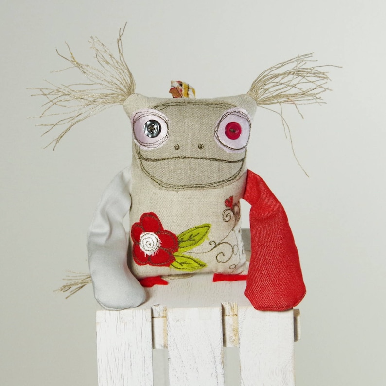 May include: A handmade plush toy with a gray linen body, red and white button eyes, and a red embroidered flower on the front. The toy has long, white yarn hair and is wearing a red fabric scarf.