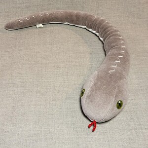 May include: A grey plush snake toy with green eyes and a red tongue. The snake is curled up in a C shape.