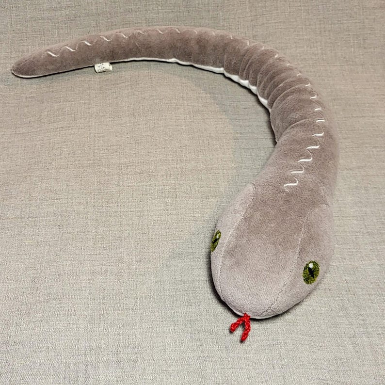 May include: A grey plush snake toy with green eyes and a red tongue. The snake is curled up in a C shape.