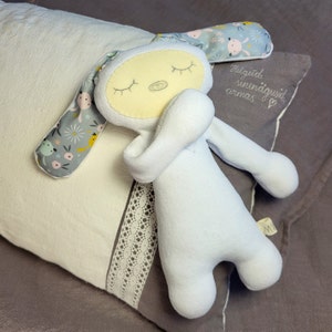 Puede incluir: Un conejito de peluche blanco con los ojos cerrados y la cara amarilla está acostado sobre una almohada blanca con un estampado floral gris y un borde de encaje. La almohada tiene el texto "Helgrid sinnaõigused armas" bordado.
