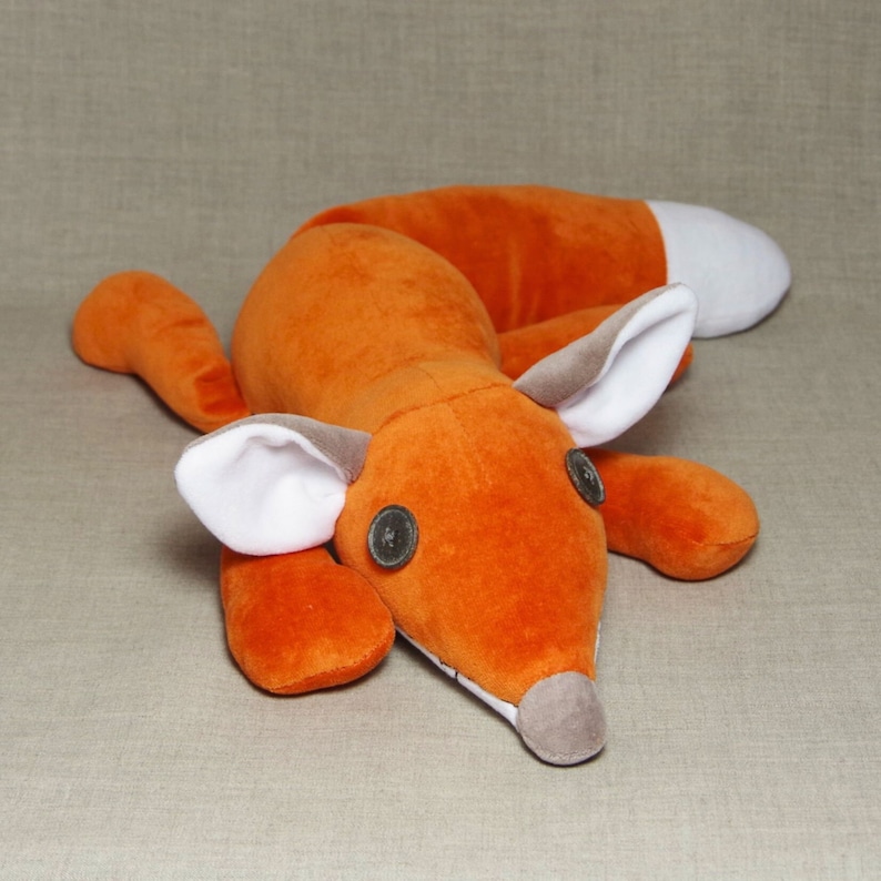 May include: A plush orange fox toy with white ears and black button eyes. The fox is lying on its belly with its head turned to the side.