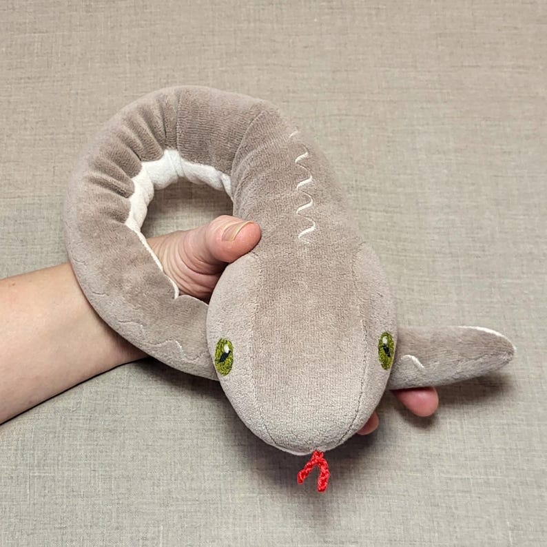 May include: A grey plush snake toy with green eyes and a red tongue. The snake is curled up in a person's hand.