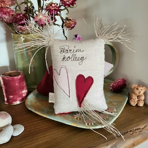 May include: A small, handmade linen pillow with the text "Parim kolleg" embroidered in brown thread. The pillow has two pink and red felt hearts sewn on the front. The pillow is sitting on a wooden surface with a green vase and dried flowers in the background.