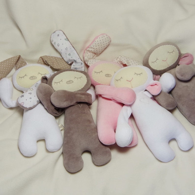 Puede incluir: Cinco conejitos de peluche en diferentes colores, incluyendo blanco, rosa, marr&oacute;n y gris. Cada conejito tiene una cara cosida con los ojos cerrados y una peque&ntilde;a nariz. Los conejitos est&aacute;n tumbados sobre una superficie blanca.