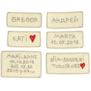 Puede incluir: Seis etiquetas de fieltro beige con texto bordado y corazones rojos. Las etiquetas dicen "Gregor", "Андрей", "Kati", "Marta 10.05.2018", "Mari-Anne 14.10.2018 kell 15.36 3210 g 47 cm" y "Siim-Sanderi Tudujanes".