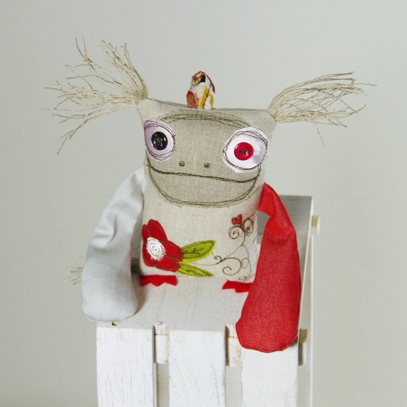 May include: A handmade plush toy with a whimsical design. The toy has a gray fabric body with embroidered details, large button eyes, and a red scarf. The toy is sitting on a white wooden chair.