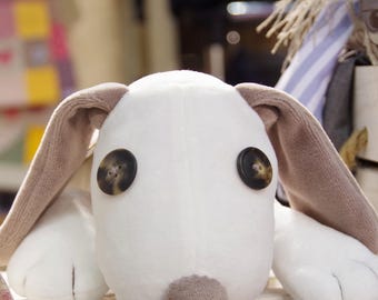 May include: A white plush dog toy with brown ears and button eyes. The dog is lying down with its head resting on its paws.