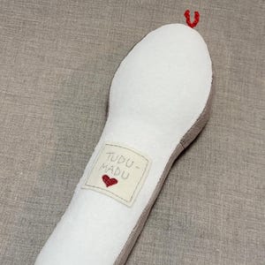 May include: A white plush toy shaped like a long, thin snake with a red heart embroidered on a tag that says "TUDDU - MADU".