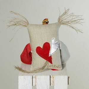 May include: A handmade linen doll with long, frayed hair. The doll is wearing a red and white heart on its chest and has a white heart on its back. The doll is sitting on a white wooden stool.