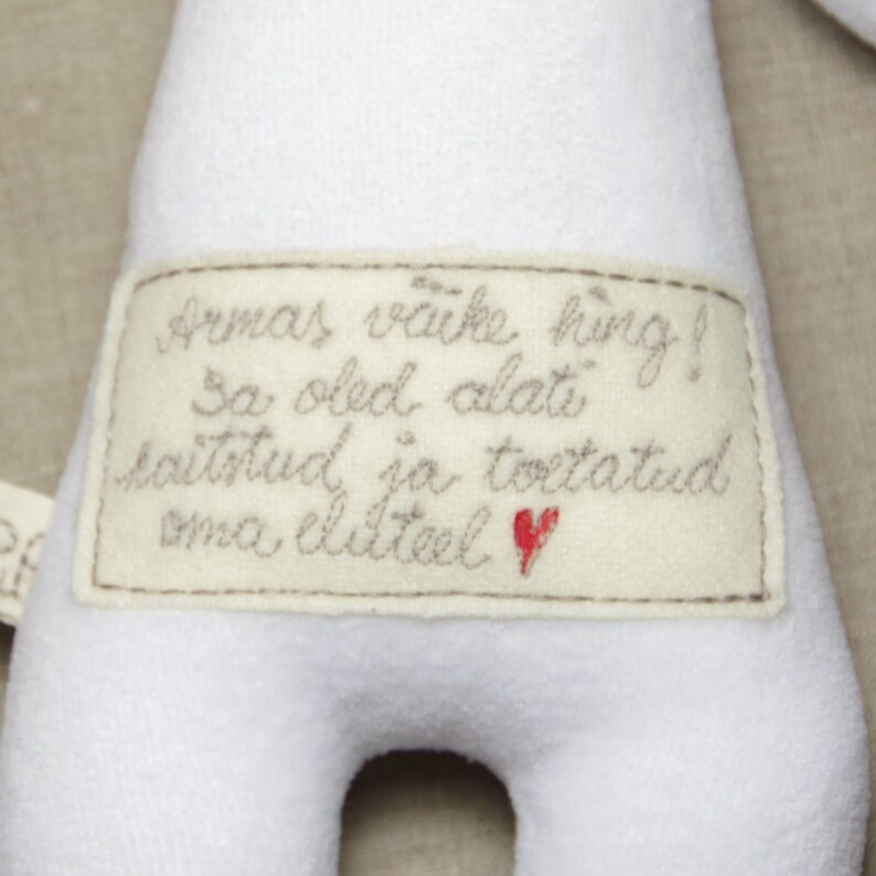 Puede incluir: Juguete de peluche blanco con una etiqueta cosida que dice "Armas v&auml;ike hing! Sa oled alati kaitstud ja toetatud oma eluteel." con un coraz&oacute;n rojo.