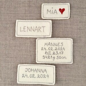 May include: Four white felt name tags with black stitching. The tags read "Mia" with a red heart, "Lennart", "Hannes 24.02.2024  kell 23.17 3421g 50cm", and "Johanna 24.02.2024".