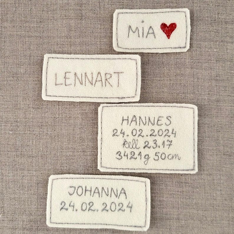 May include: Four white felt name tags with black stitching. The tags read "Mia" with a red heart, "Lennart", "Hannes 24.02.2024  kell 23.17 3421g 50cm", and "Johanna 24.02.2024".