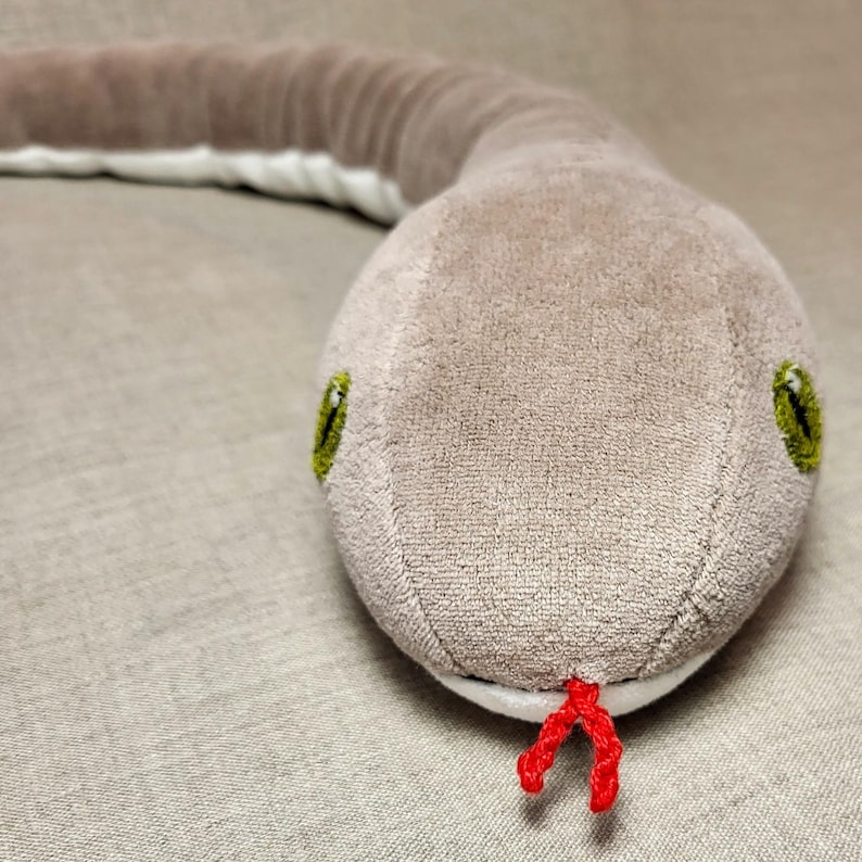 May include: A plush snake toy with a grey body, green eyes, and a red tongue. The snake is curled up on a light brown fabric surface.