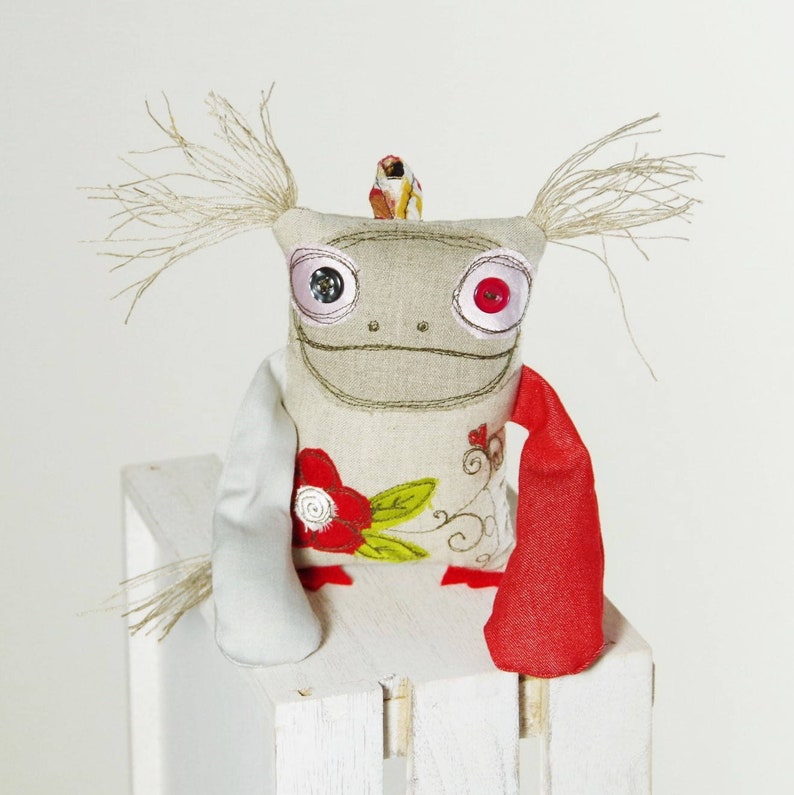 May include: A handmade plush toy with a whimsical design. It has a beige body with embroidered details, red and white buttons for eyes, and a red and white floral pattern. The toy has long, stringy hair and a small bird perched on its head.