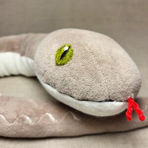 May include: A plush grey snake toy with a yellow eye and a red tongue. The snake is curled up in a circle.