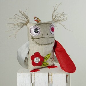 May include: A handmade plush toy with a gray linen body, red fabric arms, and a red embroidered flower. The toy has a whimsical expression with large eyes and a wide smile. The toy has a pair of long, brown yarn pigtails.