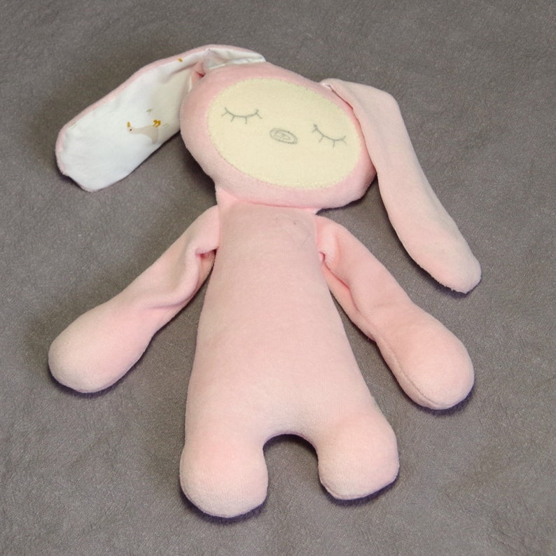 Puede incluir: Un juguete de peluche de conejo rosa con los ojos cerrados y una cara blanca. El conejo tiene orejas largas y una textura suave y mullida.