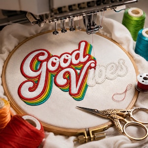 Puede incluir: Un bastidor de bordado con tela color hueso que muestra las palabras Good Vibes cosidas en hilo blanco, delineadas por anillos concéntricos de colores arcoíris. Incluye una máquina de bordar, tijeras doradas y carretes de hilo.