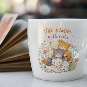 May include: White ceramic mug featuring the phrase "Life is better with cats" and a colourful illustration of five cartoon cats. The cats are in shades of white, grey, and orange, surrounded by flowers and hearts.