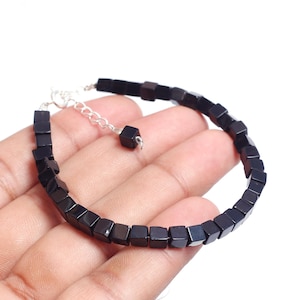 May include: A black cube bead bracelet with a silver-colored chain and clasp. The bracelet is made of small, square beads strung together. The bracelet is shown on a person's hand.