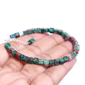 May include: A bracelet made of square-cut green and red gemstones. The beads are strung on a silver chain. The bracelet is shown against a white backdrop. The gemstones have a natural, slightly rough texture. The bracelet is approximately 18 cm in length.
