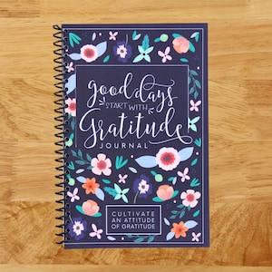May include: A spiral-bound journal with a navy blue cover featuring the words "good days start with gratitude" in white script. The cover is decorated with colorful floral and leaf illustrations. The journal also includes the text "Cultivate an attitude of gratitude".