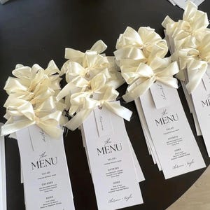 May include: White rectangular menu cards with ivory satin bows. Each card features the text "The MENU" in a modern font, with menu items listed below. The cards are arranged on a dark surface.