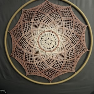 May include: A large, handmade dream catcher with a complex, geometric design. The central portion is cream-colored, surrounded by concentric circles of light brown and a web of light brown netting. The outer ring is a light tan color.