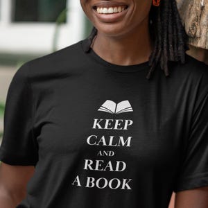 Könnte beinhalten: Schwarzes T-Shirt mit dem weißen Text "KEEP CALM AND READ A BOOK". Über dem Text befindet sich eine Grafik eines offenen Buches. Die Person lehnt an einer Backsteinmauer.