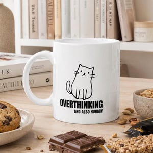 Può includere: Una tazza in ceramica bianca con un disegno a linee nere di un gatto e il testo "OVERTHINKING AND ALSO HUNGRY" stampato in nero. La tazza presenta un manico standard e una finitura liscia, con un semplice design grafico sulla superficie.