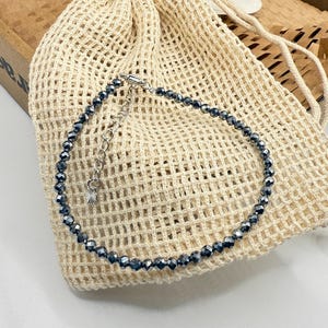May include: A dark grey beaded anklet with a silver clasp. The anklet is displayed on a beige mesh bag, with a cardboard box in the background. The anklet is a delicate piece of jewellery, suitable for everyday wear.