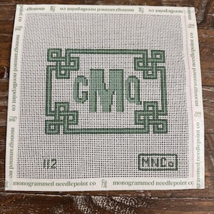 May include: A square needlepoint canvas with a green monogram design featuring the letters "CMo" within a decorative border. The canvas is white with a grid pattern and the text "monogrammed needlepoint co" is printed around the edges.