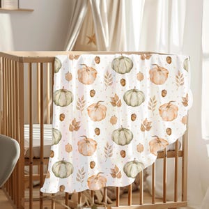 Op de afbeelding: Een witte babydeken met een herhalend patroon van aquarel pompoenen in oranje en groen, samen met bruine eikels en bladeren. De deken is over een houten wieg gedrapeerd en creëert een herfstthema.