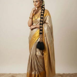 May include: A woman wearing a gold and beige sari with a gold border. The sari is draped elegantly. She is adorned with gold jewelry, including a necklace, earrings, and a headpiece. Her long, dark hair is styled with a decorative braid.