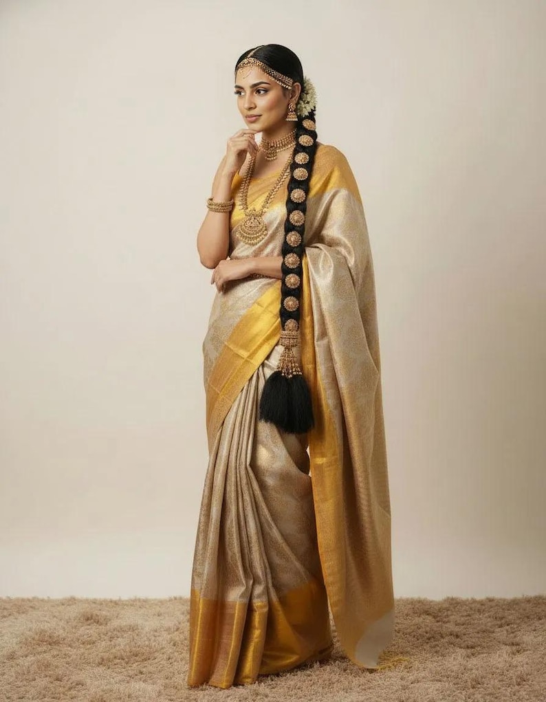 May include: A woman wearing a gold and beige sari with a gold border. The sari is draped elegantly. She is adorned with gold jewelry, including a necklace, earrings, and a headpiece. Her long, dark hair is styled with a decorative braid.