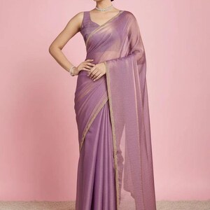 May include: A mauve-coloured saree featuring a subtle shimmering texture and a delicate gold-toned border. The garment includes a sleeveless blouse with a matching mauve hue, paired with a draped fabric that falls gracefully to the floor.
