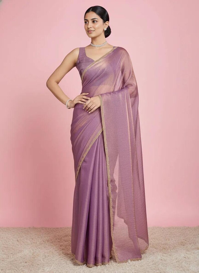May include: A mauve-coloured saree featuring a subtle shimmering texture and a delicate gold-toned border. The garment includes a sleeveless blouse with a matching mauve hue, paired with a draped fabric that falls gracefully to the floor.