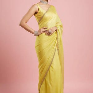 May include: A sleeveless, bright yellow sari featuring a subtle sheen and a delicate, thin embroidered border along the edges. The garment is paired with a matching sleeveless blouse, a sparkling silver-toned choker necklace, and matching bracelets.